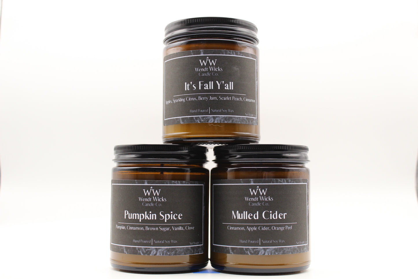 Three Wendt Wicks amber glass jar candles stacked in a pyramid with black lids. Candles labeled 'Pumpkin Spice' 'It's Fall Y'all' and 'Mulled Cider' on a white background