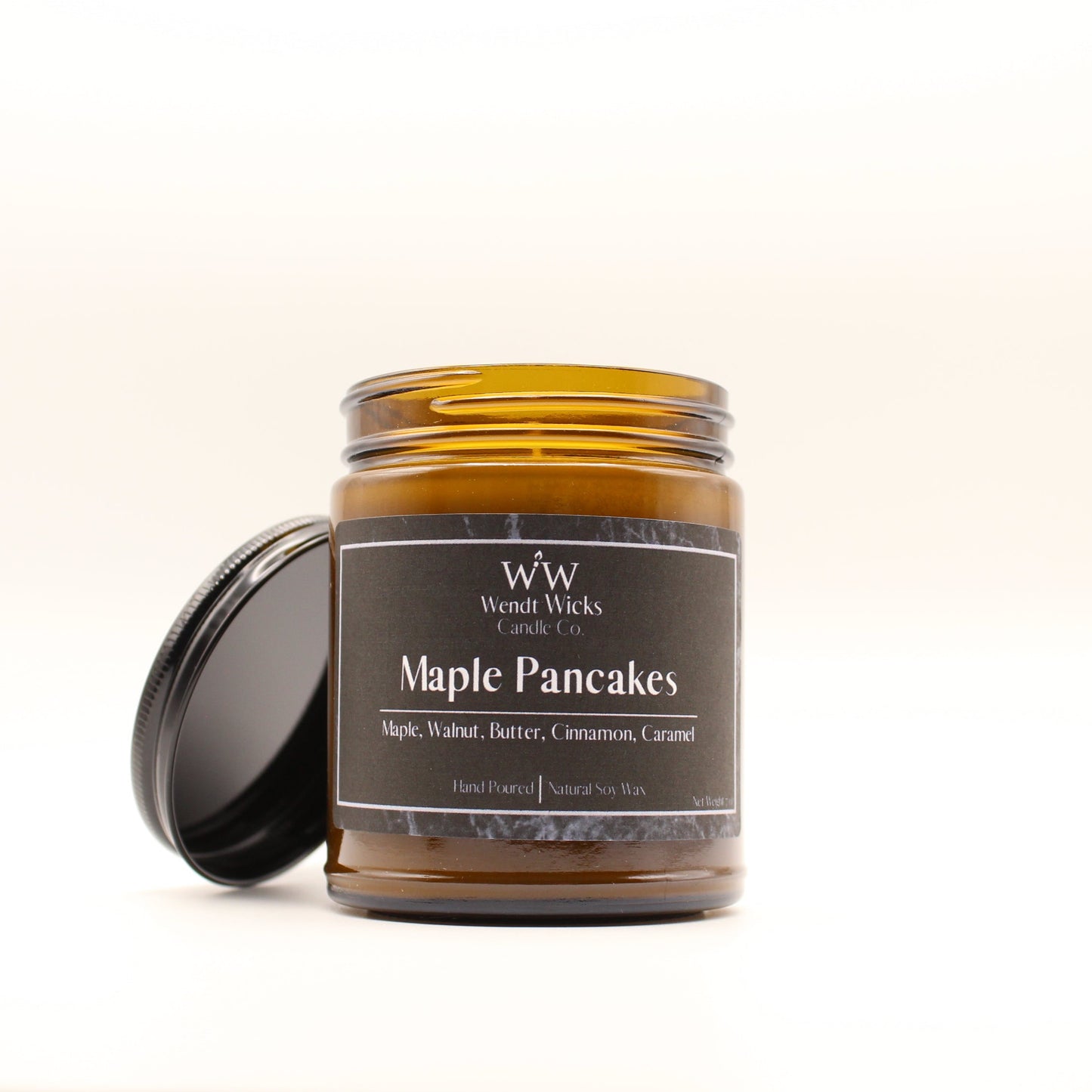 Wendt Wicks amber glass jar candle with black lid leaning on its left side. Candle labeled 'Maple Pancakes' on a white background