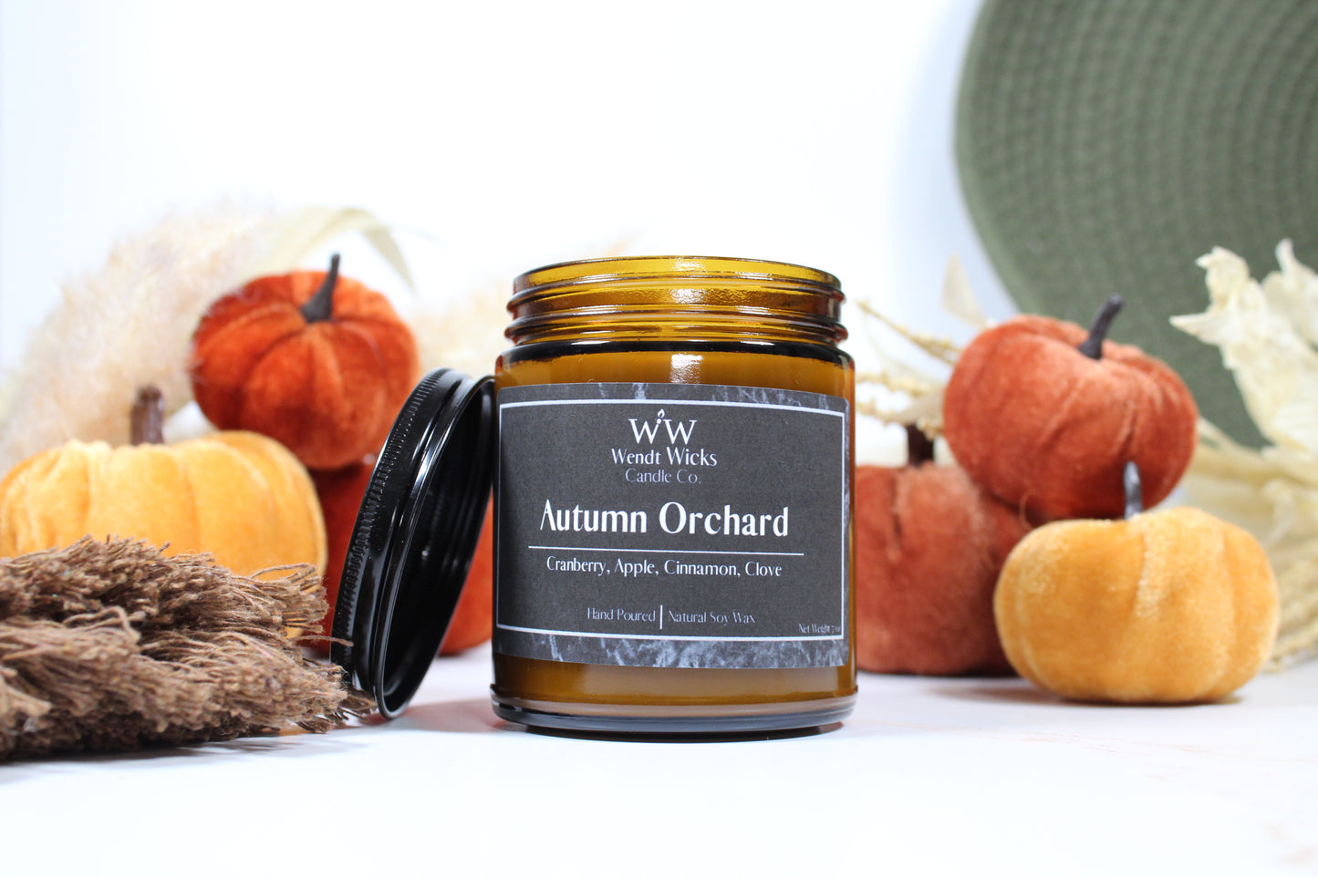 Wendt Wicks amber glass jar candle with black lid leaning against its left side. Candle labeled 'Autumn Orchard' with pumpkins and decorative Autumn elements on a white background