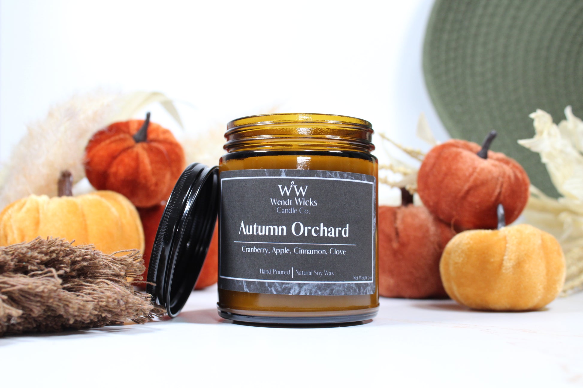 Wendt Wicks amber glass jar candle with black lid leaning against its left side. Candle labeled 'Autumn Orchard' with pumpkins and decorative Autumn elements on a white background