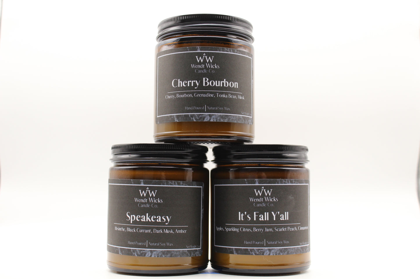 Three Wendt Wicks amber glass jar candles stacked in a pyramid with black lids. Candles labeled 'Speakeasy' 'It's Fall Y'all' and 'Cherry Bourbon' on a white background