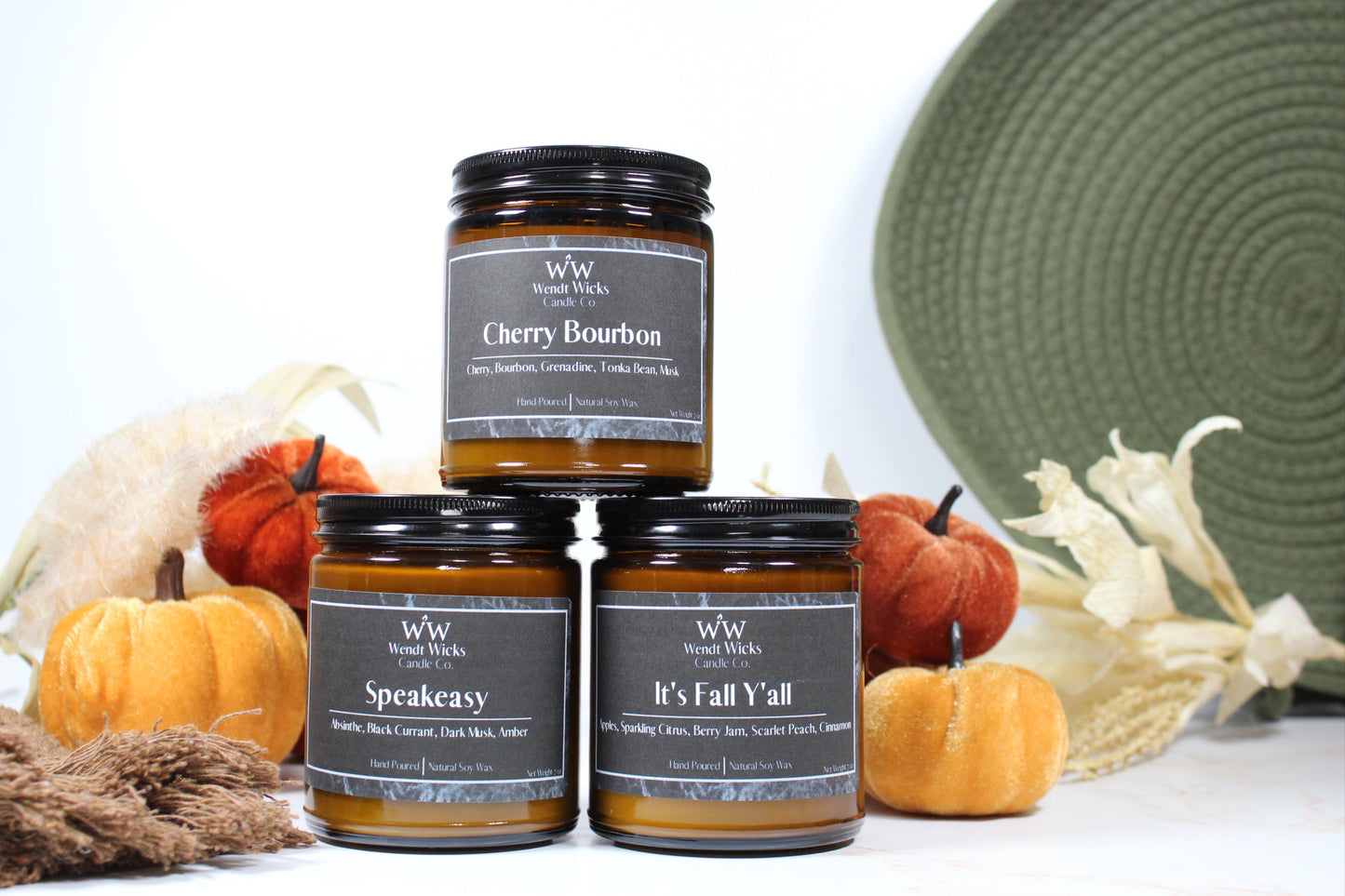 Three Wendt Wicks amber glass jar candles stacked in a pyramid with black lids. Candles labeled 'Speakeasy' 'It's Fall Y'all' and 'Cherry Bourbon' with pumpkins and decorative Autumn elements on a white background