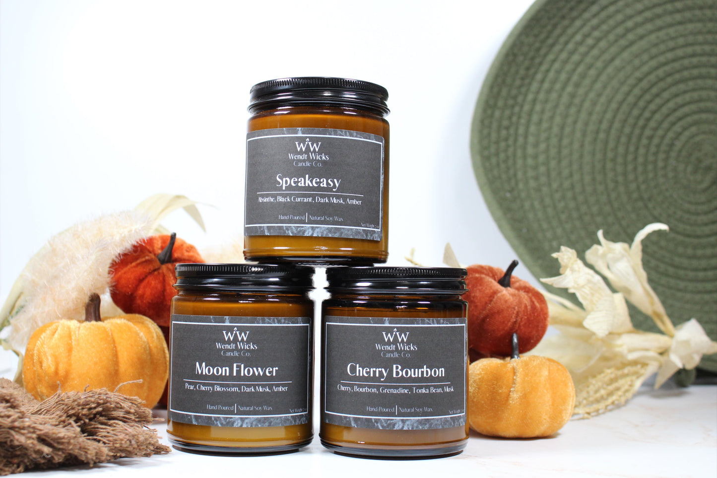 Three Wendt Wicks amber glass jar candles stacked in a pyramid with black lids. Candles labeled 'Speakeasy' 'Moon Flower' and 'Cherry Bourbon' with pumpkins and decorative Autumn elements on a white background