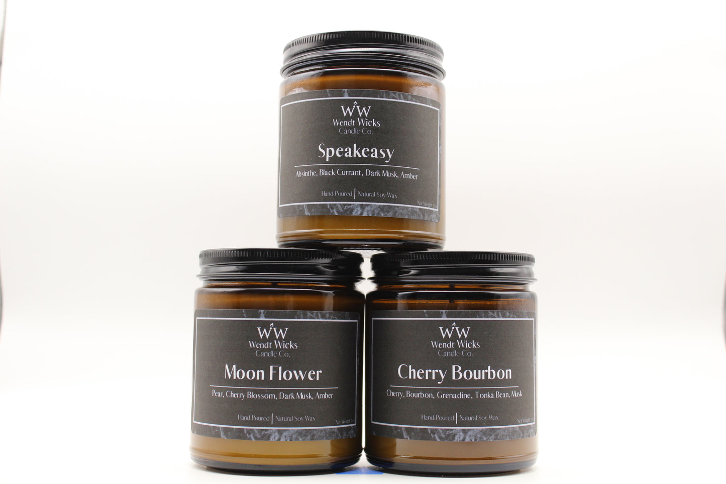 Three Wendt Wicks amber glass jar candles stacked in a pyramid with black lids. Candles labeled 'Speakeasy' 'Moon Flower' and 'Cherry Bourbon' on a white background