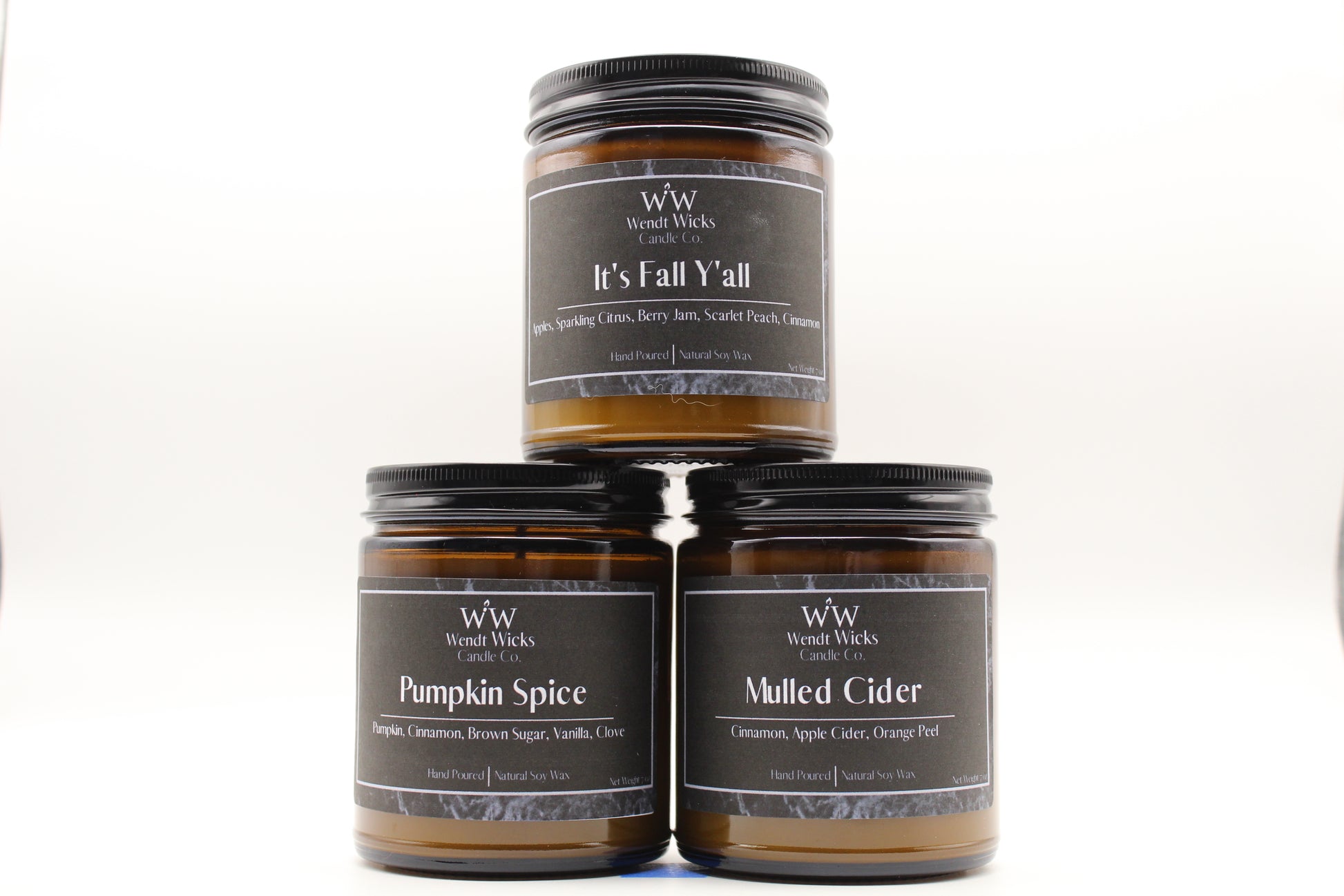 Three Wendt Wicks amber glass jar candles stacked in a pyramid with black lids. Candles labeled 'Pumpkin Spice' 'It's Fall Y'all' and 'Mulled Cider' on a white background