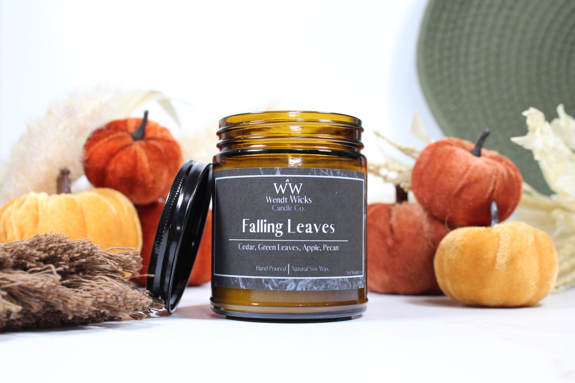 Wendt Wicks amber glass jar candle with black lid leaning on its left side. Candle labeled 'Falling Leaves' with pumpkins and decorative Autumn elements on a white background