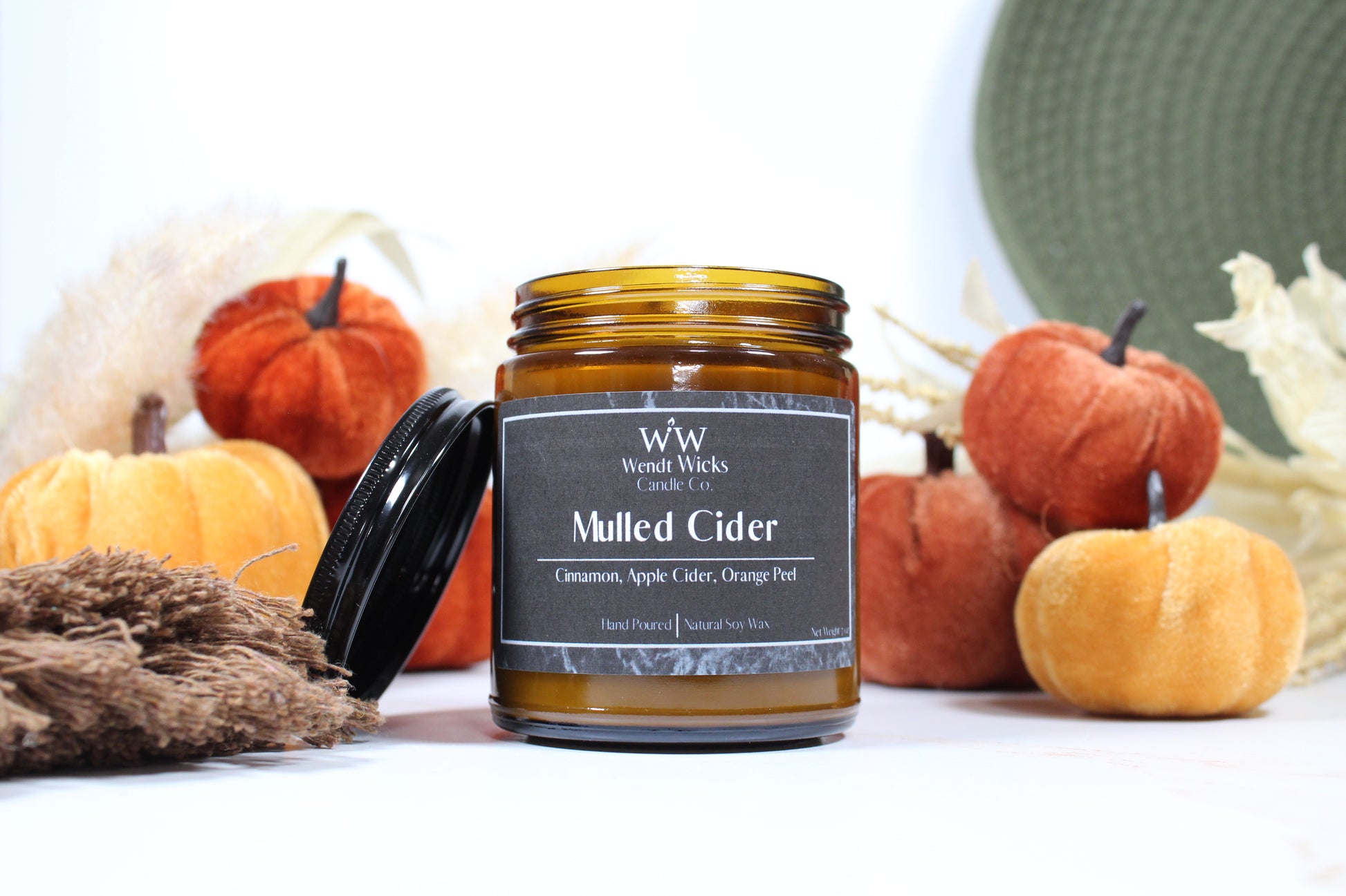 Wendt Wicks amber glass jar candle with black lid leaning against its left side. Candle labeled 'Mulled Cider' with pumpkins and decorative Autumn elements on a white background