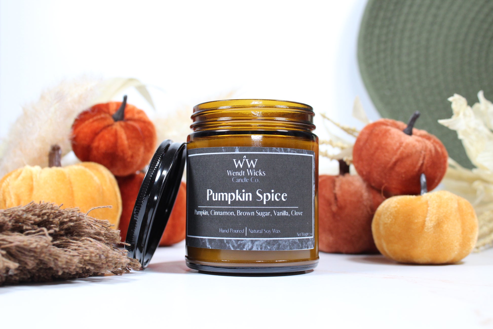 Wendt Wicks amber glass jar candle with black lid leaning against its left side. Candle labeled 'Pumpkin Spice' with pumpkins and decorative Autumn elements on a white background